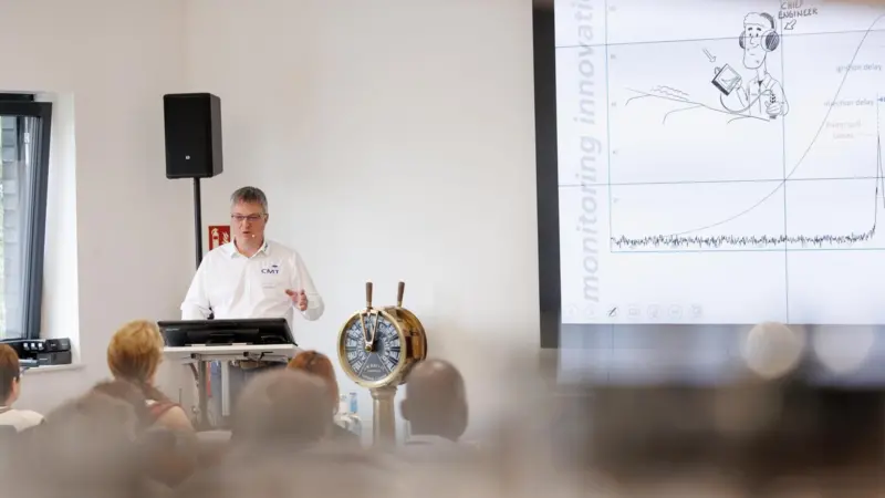 A trainer stands by a white board and a ship's wheel training people